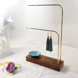 May include: A gold metal earring stand with two tiers and a wooden base. The stand is holding a pair of teal tassel earrings.