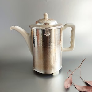Könnte beinhalten: Eine silberne, gehämmerte Kaffeekanne mit einem weißen Porzellanhenkel und -deckel. Die Kanne hat einen runden Boden mit drei Füßen.