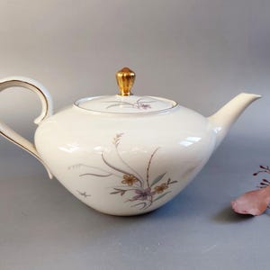 May include: White porcelain teapot with a gold-colored handle and lid finial. The teapot features a floral design in muted colors. The spout and handle are curved. A classic piece for serving tea.