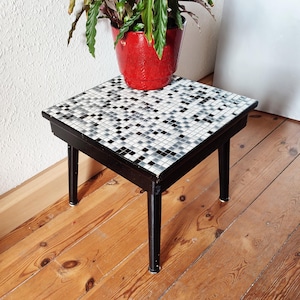 May include: A black side table with a black and white checkered tile top. The table has three legs and is sitting on a wooden floor.