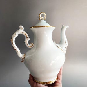 Puede incluir: Una tetera de porcelana blanca con adornos y detalles dorados. La tetera tiene un asa y un pico curvos, y una tapa con un adorno dorado. El diseño es ornamentado y elegante, adecuado para servir té.