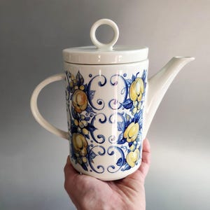 Può includere: Teiera in ceramica color crema con coperchio e manico. Decorata con un motivo floreale blu e giallo. Il beccuccio è lungo e sottile e il manico è curvo. Per servire il tè.