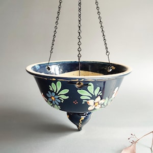 May include: A hanging ceramic planter with a dark blue glaze and floral designs in green, blue, and white. The planter is suspended by black chains and has a decorative, rounded base. The rim is a lighter color.