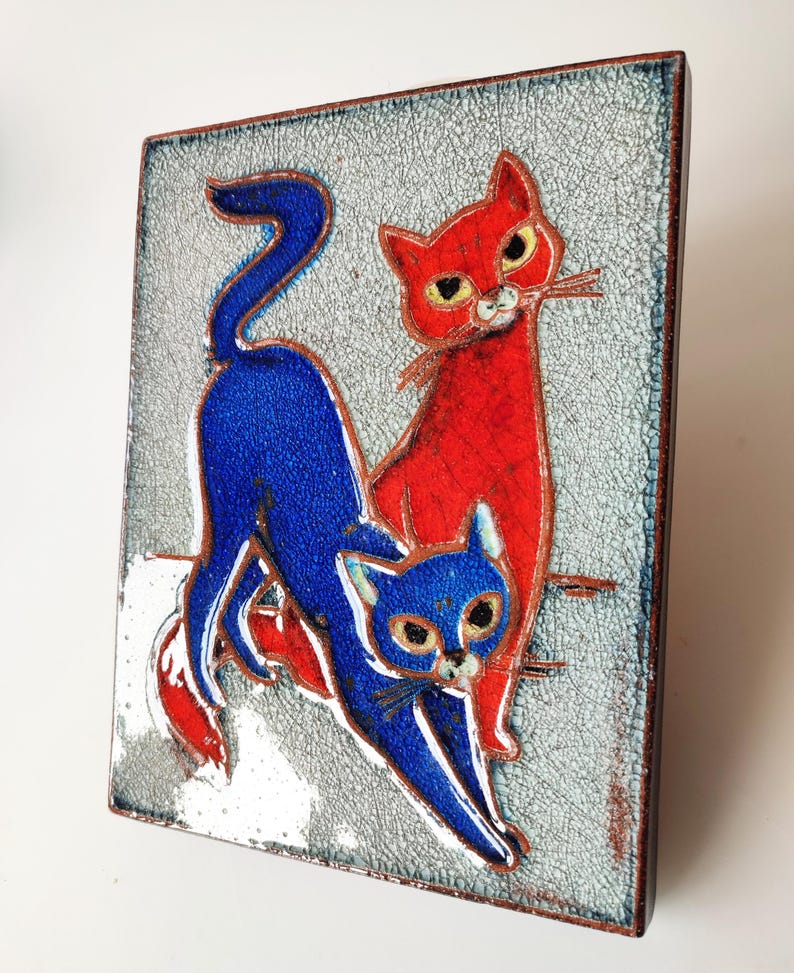 May include: A rectangular ceramic tile featuring two stylized cats. One cat is red, the other is blue, set against a textured, light gray background. The tile has a dark brown border and is likely a decorative piece.