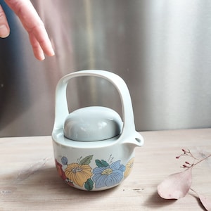 May include: A light blue ceramic teapot with a floral design. The teapot has a lid and a handle. The flowers are yellow, blue, and red.