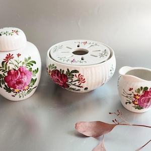 May include: A white ceramic tea set featuring a lidded jar, a round warmer with a perforated lid, and a small creamer. Each piece is decorated with a large pink rose and green leaves, with red and yellow accents. The set is designed for serving tea.