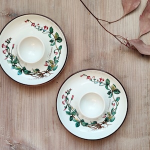 Może przedstawiać: Dwa białe ceramiczne stojaki na jajka z brązową obwódką i wzorem kwiatowym. Wzór przedstawia różowe kwiaty i zielone liście na białym tle.