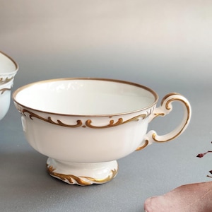 Op de afbeelding: Witte theekop met gouden rand en een decoratief oor. De kop heeft een voetstuk en gouden details rond de rand en de basis. Onderdeel van een theeservies.