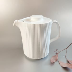 May include: A white ceramic teapot with a lid and handle. The body of the teapot has vertical ribbed detailing. The teapot is on a light grey surface. A sprig of dried leaves is next to the teapot.