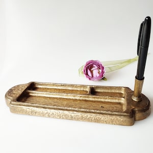 Op de afbeelding: Een gouden metalen bureau-organizer met drie vakken en een pennenhouder. De organizer heeft een getextureerd oppervlak en een decoratief ontwerp. Een zwarte pen zit in de houder.