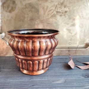 Puede incluir: Un macetero de cobre con un diseño acanalado. El macetero está vacío y se encuentra sobre una superficie de madera.