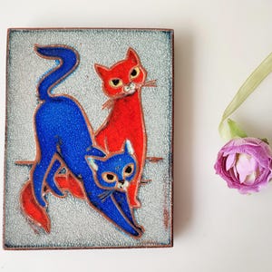 May include: A rectangular ceramic tile featuring two stylized cats. One cat is blue, stretching, and the other is red, sitting upright. The tile has a textured, light gray background and a dark brown border. A purple flower and green ribbon are in the background.