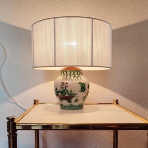 May include: A table lamp with a white linen shade and a ceramic base with green and red floral designs.
