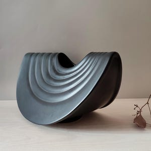 Pode incluir: Uma tigela decorativa de cerâmica cinza escuro com um design curvo único. A tigela apresenta uma série de cristas em forma de onda no exterior. O interior é preto e a tigela assenta numa superfície clara.