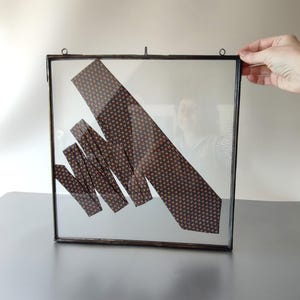 May include: A framed necktie with a repeating pattern of small brown and tan shapes. The tie is folded and arranged diagonally within a clear glass frame with a dark metal border. The frame has a small hanging loop.