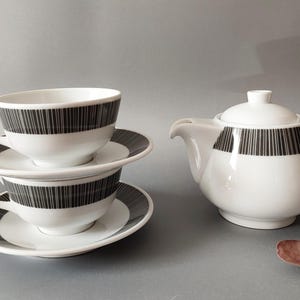 Może przedstawiać: Biały ceramiczny zestaw do herbaty z czarnym wzorem w pionowe paski. Zestaw zawiera dwie filiżanki do herbaty na spodkach i czajniczek z pokrywką. Filiżanki i czajniczek mają pasujący czarny wzór w paski pośrodku.