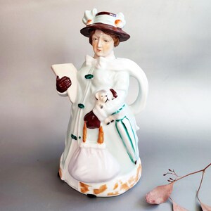 Może przedstawiać: Ceramiczny dzbanek do herbaty w kształcie postaci w brązowym kapeluszu i jasnoniebieskim płaszczu. Figurka trzyma książkę, małego białego psa i torebkę, a pod ramieniem ma zielony parasol. Podstawa ozdobiona jest brązowym, cętkowanym wzorem.