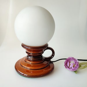 May include: A brown ceramic table lamp with a white globe shade. The lamp has a unique design with a handle on the side. The base of the lamp is decorated with brown and white swirls.