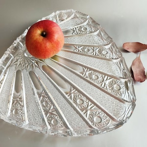 Può includere: Un piatto da portata in vetro trasparente tagliato con un design a ventaglio, contenente una mela rossa. Il piatto presenta intricati motivi geometrici e un bordo smerlato. Foglie secche a lato.