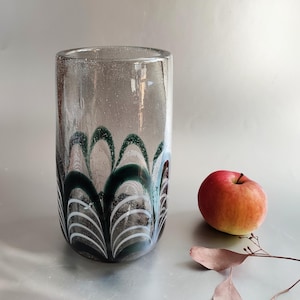 Könnte beinhalten: Eine klare Glasvase mit einem dekorativen Muster aus dunkelgrünen und weißen geschwungenen Linien. Die Vase ist zylindrisch. Ein roter Apfel liegt neben der Vase. Die Vase ist etwa 20 cm hoch.
