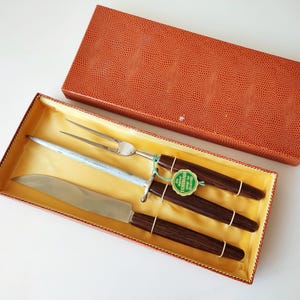 Puede incluir: Un juego de trinchar vintage con cuchillo, tenedor y afilador, con mangos de madera oscura y una caja naranja decorativa. La caja tiene un patrón texturizado. El juego incluye una etiqueta verde.