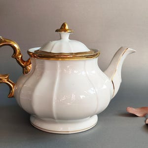 Könnte beinhalten: Eine weiße Porzellan-Teekanne mit goldenen Akzenten. Die Teekanne hat einen geschwungenen Griff und Ausguss, einen goldverzierten Deckel und einen goldenen Knauf. Der Körper der Teekanne hat ein geripptes Design. Ein dekoratives Stück zum Servieren von Tee.
