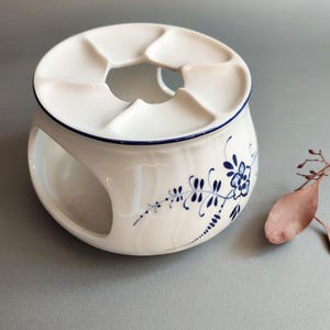 以下が含まれることがあります： 青い花柄の白いセラミックティーウォーマー。ウォーマーは、円形の開口部のある丸いベースと、複数の開口部のある上部を備えています。青い線が上端をトリミングしています。