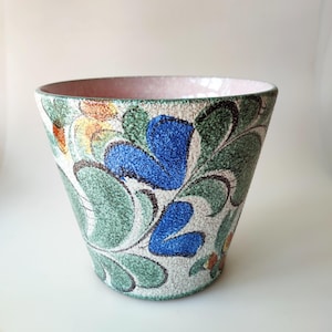 Puede incluir: Una maceta de cerámica con fondo blanco y un patrón floral en tonos azules, verdes, amarillos y naranjas. La maceta tiene un borde rosa y una superficie texturizada.