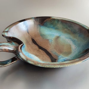 May include: A ceramic serving dish with a handle, featuring a blue, brown, and black glaze. The dish has a unique, organic shape and a glossy finish.