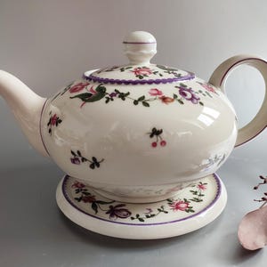 Può includere: Teiera in ceramica color crema con coperchio e piattino abbinati. La teiera e il piattino sono decorati con motivi floreali rosa, viola e verdi, delineati con bordi viola. La teiera ha un beccuccio curvo e un manico arrotondato.