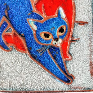 May include: A square ceramic tile featuring a stylized blue cat with orange accents against a textured, light gray background. The cat has large eyes and is outlined in a dark brown color. The tile has a crackle finish.