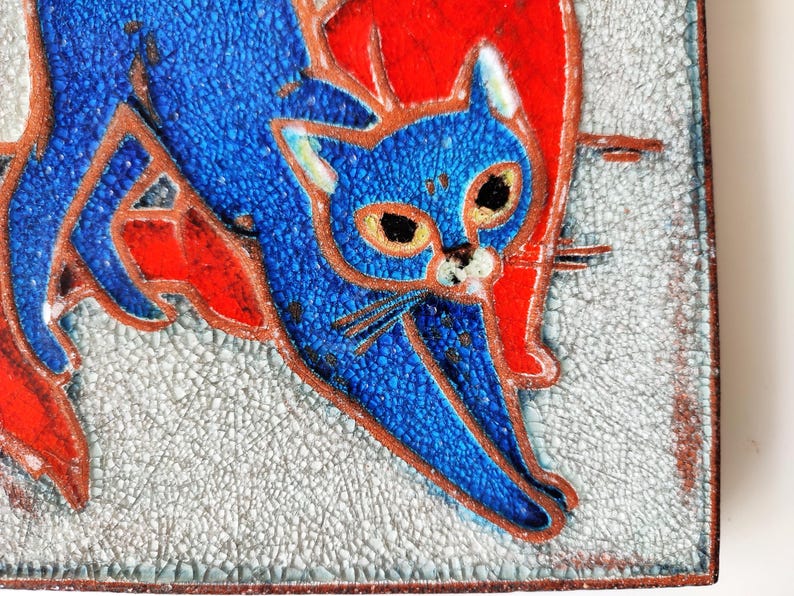May include: A square ceramic tile featuring a stylized blue cat with orange accents against a textured, light gray background. The cat has large eyes and is outlined in a dark brown color. The tile has a crackle finish.
