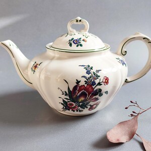 May include: Cream-colored ceramic teapot with a floral design. The teapot features a curved spout, a handle, and a lid with a small handle. The floral design includes red, blue, and yellow flowers. The teapot is on a light gray surface.