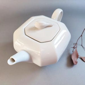 Puede incluir: Una tetera de cerámica blanca con un diseño geométrico. La tetera tiene un pico, un asa y una tapa con un pequeño pomo. La tetera está sobre una superficie gris.