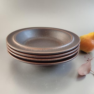 May include: A stack of four brown ceramic plates with a textured surface and a copper-colored rim. A yellow pear and a sprig of dried leaves are next to the plates. The plates have a shallow, bowl-like design.