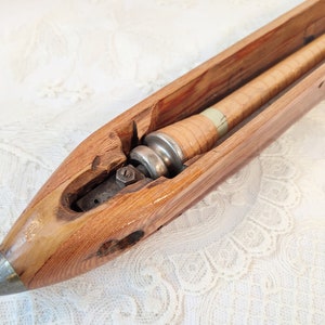 May include: A vintage wooden weaving shuttle with a metal tip and internal components. The shuttle is made of light brown wood and has a cylindrical component wrapped in tan material. The metal tip is silver.