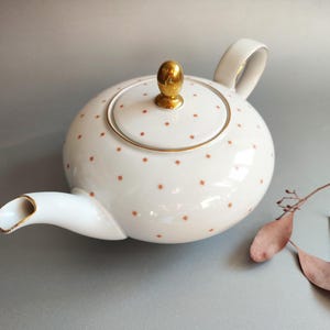 Puede incluir: Tetera de cerámica blanca con cuerpo redondeado, adornada con pequeños patrones en forma de estrella naranja. La tapa tiene un pomo dorado, y el pico y el asa tienen ribetes dorados. Un artículo decorativo para los amantes del té.