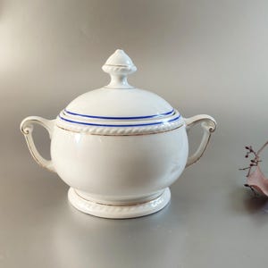 Puede incluir: Azucarero de porcelana blanca con tapa y dos asas. El tazón tiene un borde azul y dorado alrededor de la parte superior.