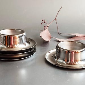 Puede incluir: Tazas de huevo plateadas brillantes con platillos a juego. Se ve una pila de tres tazas y platillos, junto con dos tazas y platillos individuales. Una ramita de hojas secas y una sola hoja están en el fondo. Las tazas tienen una superficie reflectante.