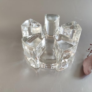 Könnte beinhalten: Ein klarer Glaskerzenhalter mit fünf einzelnen Kerzenhaltern. Der Halter hat ein strukturiertes, facettiertes Design und eine kreisförmige Basis. Auf einem der Kerzenhalter ist ein Aufkleber zu sehen. Der Hintergrund ist eine hellgraue Oberfläche.