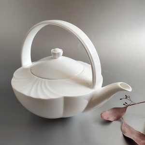 May include: White porcelain teapot with a unique curved handle and a lid. The teapot has a smooth, white finish and a delicate design.