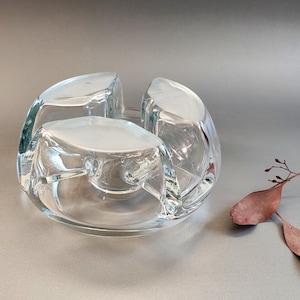 Op de afbeelding: Transparante glazen asbak met een uniek, afgerond ontwerp. De asbak heeft drie verhoogde secties en een cirkelvormige basis. Het transparante glas laat licht door. Een decoratief item voor een huis of kantoor.