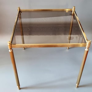 Puede incluir: Una mesa auxiliar vintage con una tapa rectangular de vidrio ahumado y un marco de metal color latón. Las patas son estriadas y rematadas con pináculos decorativos. La mesa mide aproximadamente 51 cm de largo, 51 cm de ancho y 51 cm de alto.