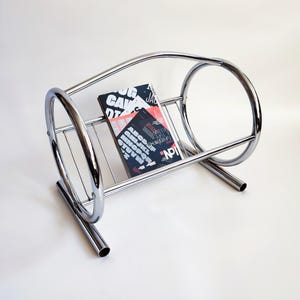 May include: A chrome magazine rack with a curved design. The rack holds several magazines with colorful covers and text. The rack has a minimalist, modern aesthetic and is set against a white background.