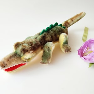 May include: A plush crocodile toy with a green back, brown and yellow body, and red teeth. The toy is lying on a white surface.