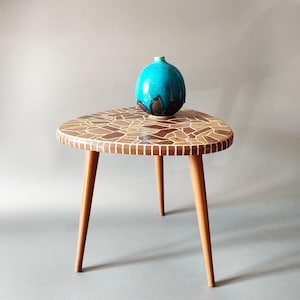 Könnte beinhalten: Ein Beistelltisch im Vintage-Stil mit einer Mosaik-Tischplatte und drei Holzbeinen. Auf dem Tisch steht eine türkisfarbene Keramikvase mit dunkelbraunem Sockel. Die Tischplatte ist ein Mosaik aus braunen, beigen und cremefarbenen Fliesen.