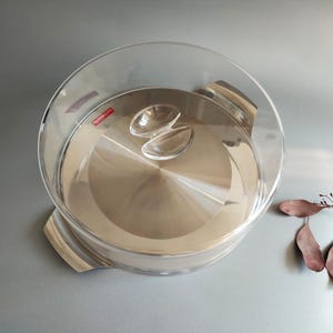 May include: A round serving dish with a clear glass dome lid and a polished stainless steel base. The lid features a clear glass handle on top. A small red rectangular label with white text reading "Techni-com" is visible on the glass lid.