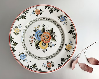May include: A white ceramic dinner plate with a floral design. The plate features a central motif of blue, orange, and yellow flowers with green leaves, surrounded by a border of smaller floral patterns in shades of blue, yellow, and orange.