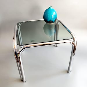 Peut inclure: Une table d'appoint carrée dotée d'un cadre en métal chromé réfléchissant et d'un plateau en verre gris fumé. Sur le plateau repose un petit vase en céramique ronde avec un émail bleu turquoise vif passant à une base noire foncée.