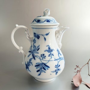 Puede incluir: Una jarra de crema de porcelana blanca con un patrón floral azul y blanco. La jarra tiene una tapa con un pequeño pomo en la parte superior.